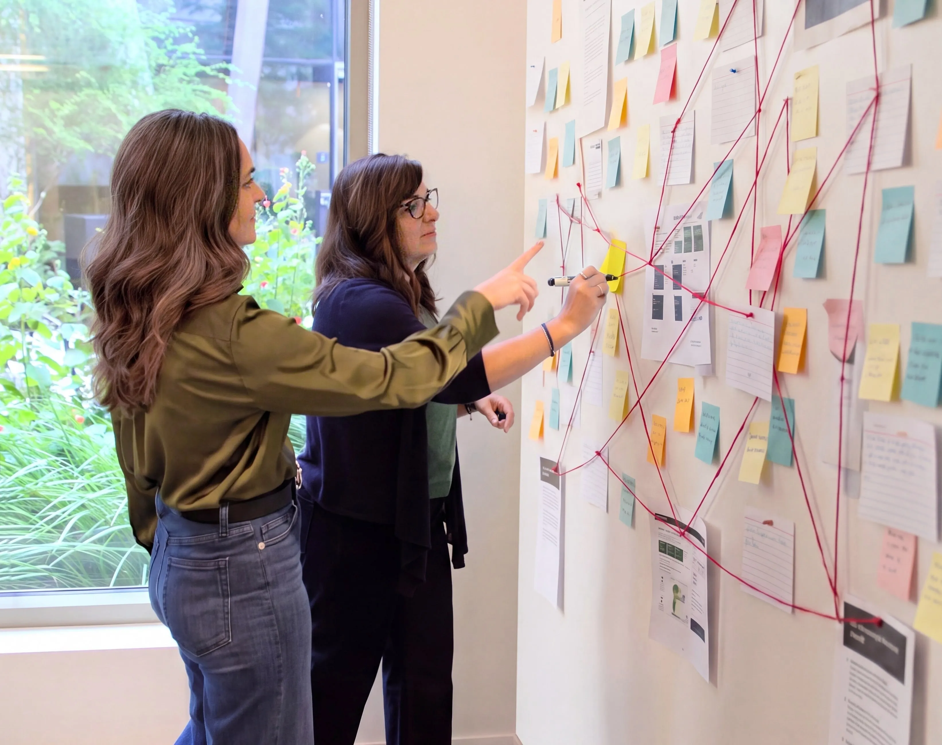 Regina Raudabaugh and Carrie Quintanilla working together at a wall of sticky notes and printouts strung together with red thread