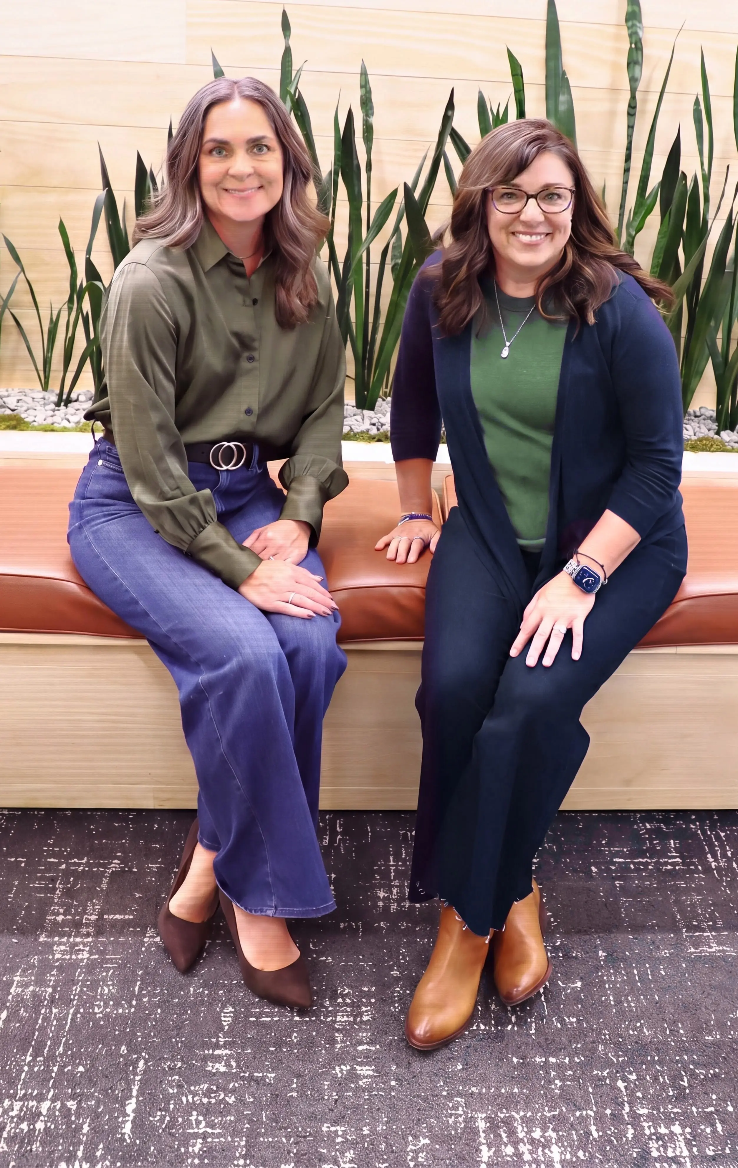 Regina Raudabaugh and Carrie Quintanilla sitting side by side on a leather bench in front of a planter of tall green plants, smiling at the camera